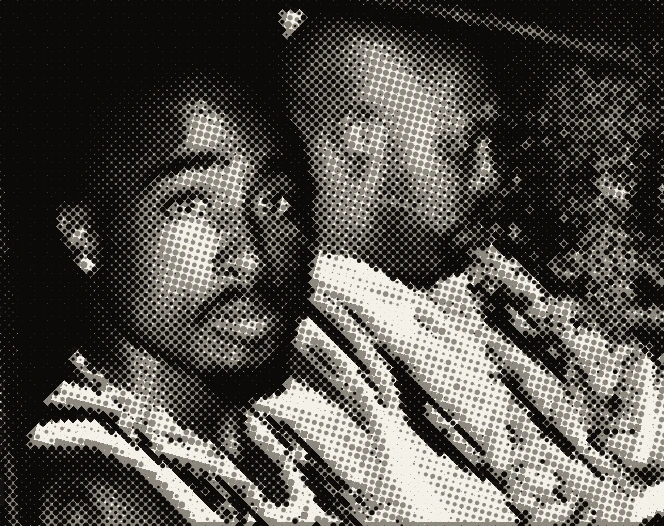 Tupac in the passenger seat of a car, next to him a bald man at the wheel.